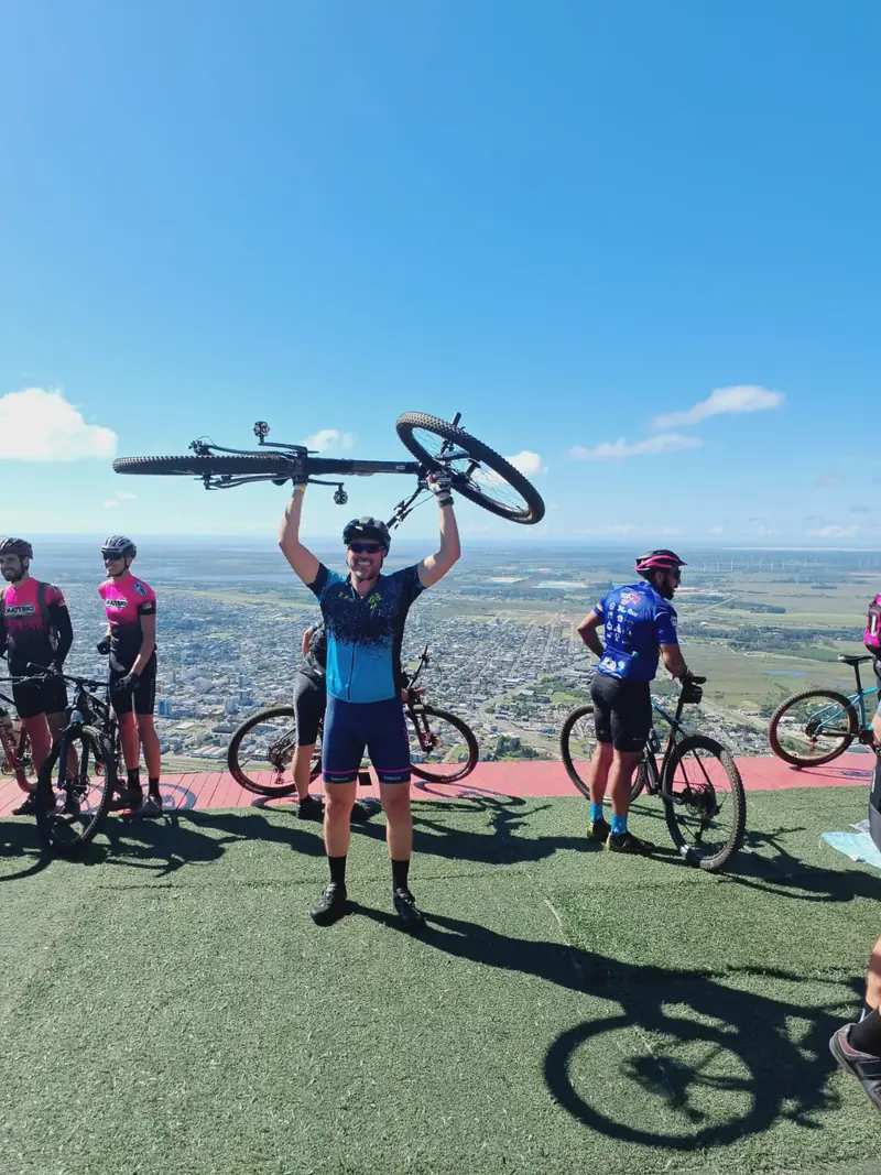 Ciclista erguendo a bike acima da cabeça no alto de um morro, com a cidade ao fundo