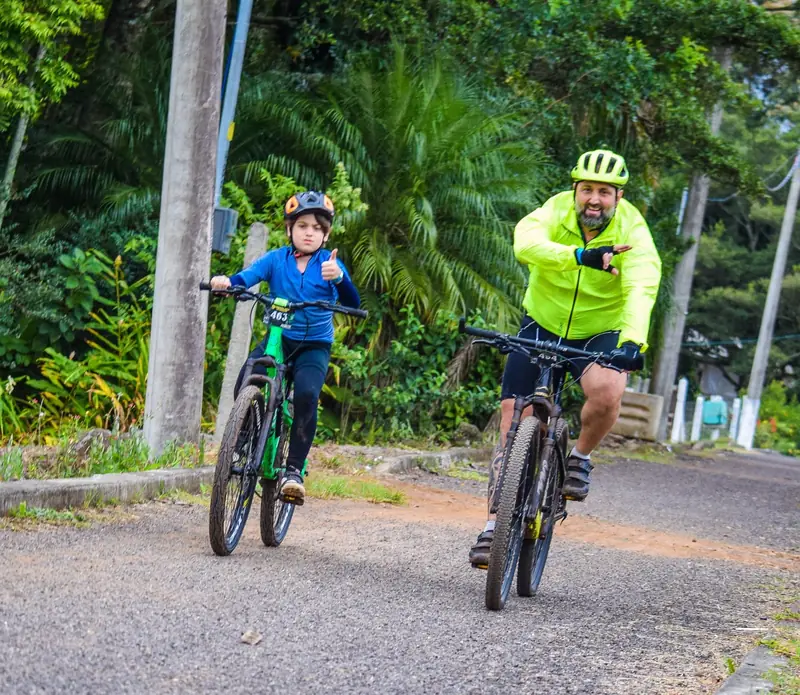 Adulto e criança pedalando lado a lado em rua arborizada durante o passeio