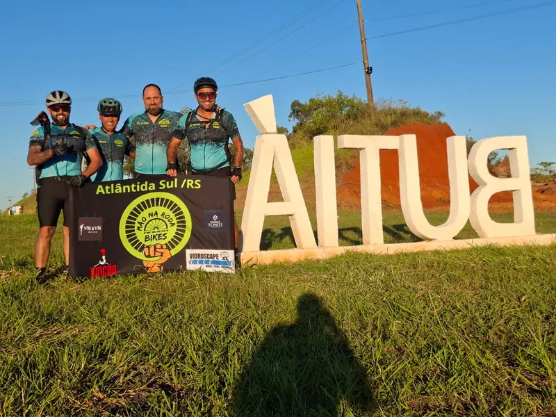 Ciclistas posam com a faixa da Mão na Roda Bikes de Atlântida Sul diante de um letreiro na estrada