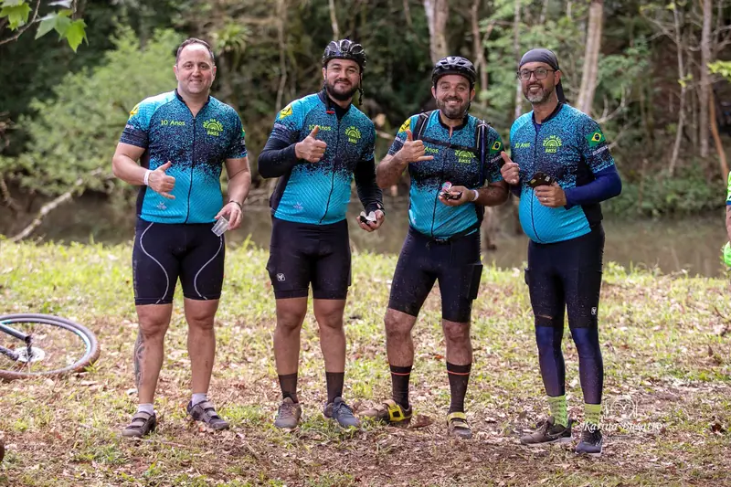 Quatro ciclistas de uniforme azul e verde posam na mata fazendo sinal de positivo