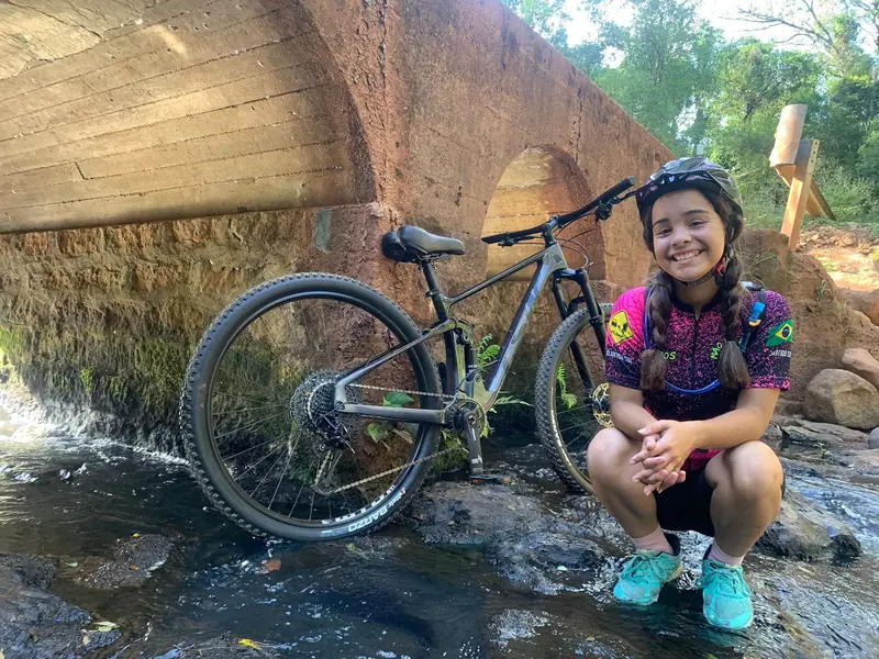 Ciclista sorrindo ao lado da bike, sobre as pedras de um riacho, junto a uma ponte de pedra
