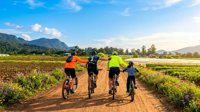 Quatro ciclistas, adultos e uma criança, pedalando lado a lado em estrada de terra entre canteiros de flores, com morros ao fundo